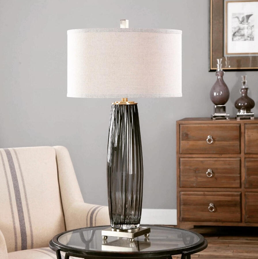 The beautiful Vilminore Table Lamp. Grooved, transparent charcoal gray glass, accented with antiqued brass plated details. The drum shade is light beige with natural stubbing. Dimensions: 33H Shade 10Hx17 Dia.